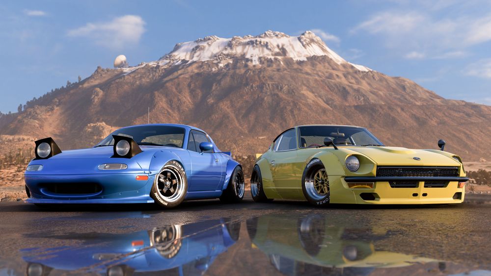 Two Rocket Bunny cars, one blue, the other yellow, parked sideways with reflections underneath in front of the Gran Caldera volcano.