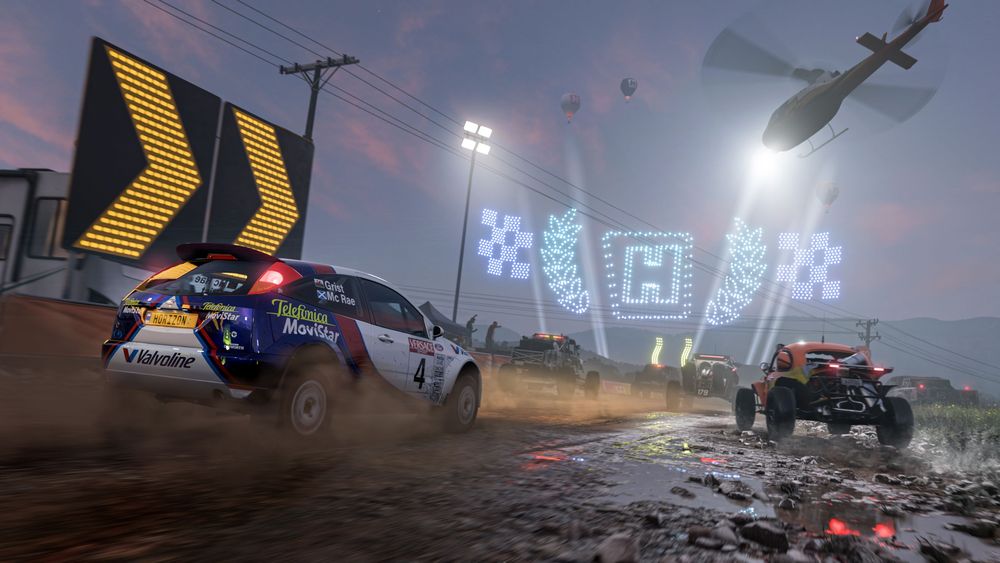 Colin McRae's Ford Focus RS kicks up the mud alongside the orange Hoonigan Baja Beetle as they race towards the Horizon Festival's drone lighting show under the spotlight of the Horizon Rally Helicopter at night time.