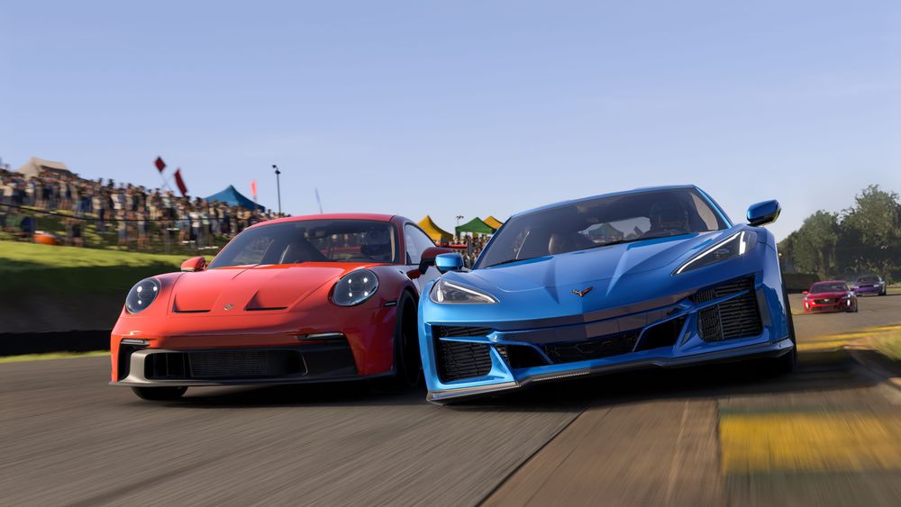 Two road-going sports cars battling for position at a circuit.  The cars are facing toward the camera with a red Porsche 911 on the left side of the image and a blue Corvette on the right side of the image, it is day time with clear skies and a helicopter