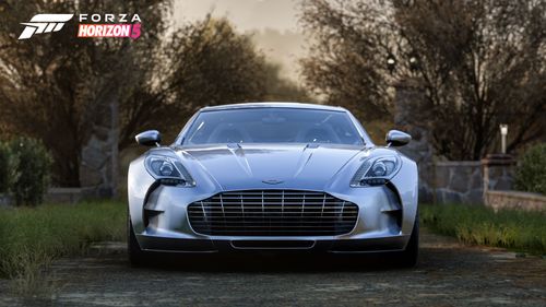 A silver 2010 Aston Martin One-77 parked at a path surrounded by grass and light trees.