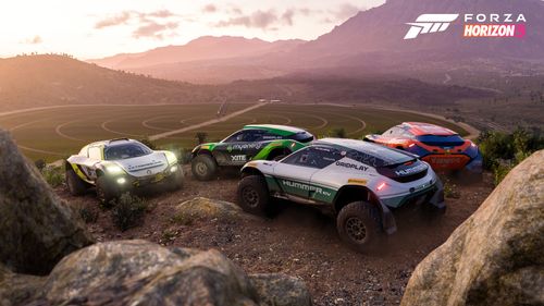 Four Extreme E off-road race cars parked by rocks take in a stunning view of Mexico's farmland and surrounding mountains at sunset.