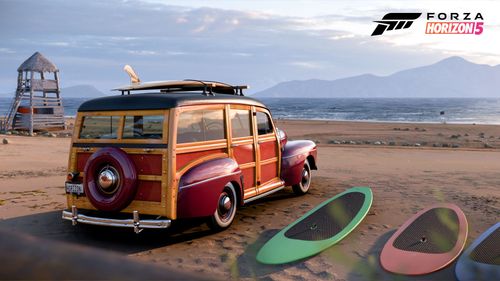 A Ford Super Deluxe Station Wagon is parked next to surfboards on the beach.