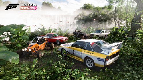 A group of rally cars parked in the rainforest including an orange buggy, a red half-truck and a white and yellow rally car. A waterfall and tall forest trees can be seen in the background.