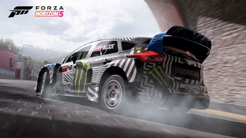 A Hoonigan GYMKHANA 9 Ford Focus RS RX with a black, white and blue livery featuring a green Monster Energy logo lifts some tire smoke drifting under a tunnel in the city of Guanajuato.