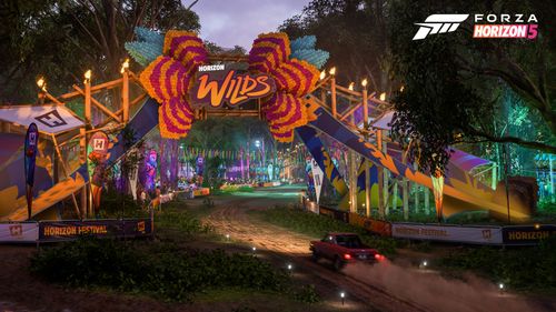 Entrance to the redecorated Horizon Wilds Festival. The scene is at night-time with orange and green lights visible. A car drives up a dust trail as it drives into the outpost.