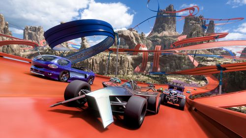 Several Hot Wheels vehicles including a purple Deora and purple Baja Boneshaker are led by a silver Bad to the Blade single-seater race car. They drive together towards the camera while surrounded by a web of blue and orange tracks in the canyon biome.
