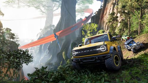 A yellow off-road Ford Bronco with its wheels turned approaches the camera as other cars follow in a dense jungle environment filled with trees and shrubbery. Two tall trees are surrounded by a large, twisting orange track. 