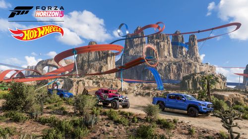 A trio of cars including a blue Ford Raptor, a pink Hot Wheels Baja Boneshaker, and a blue Subaru drive along the canyon. Large rock structures surrounded by a web of blue and orange tracks occupy the background. There is also visible foliage, ramps, and 