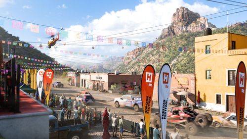 Several cars and buggies drive through the town of Pueblo Artza with orange and white Horizon Festival race flags, as well as orange, yellow and white buildings, rocky terrain in the background, and a helicopter in the sky.