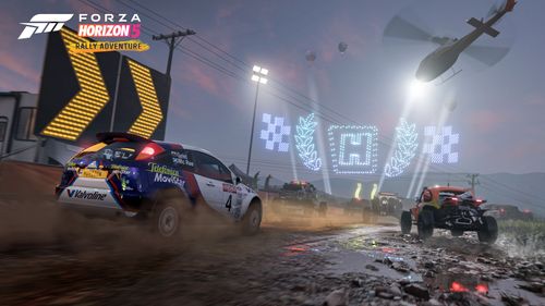 Colin McRae's Ford Focus RS kicks up the mud alongside the orange Hoonigan Baja Beetle as they race towards the Horizon Festival's drone lighting show under the spotlight of the Horizon Rally Helicopter at night time.