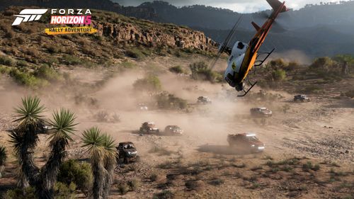 Several vehicles kick up dust as they drive through rugged terrain with the white and orange Horizon Rally Helicopter spectating the action from above.