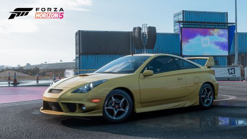 A yellow 2003 Toyota Celica SS-I parked by the Street Scene festival outpost with shipping containers and an animated screen in the background.