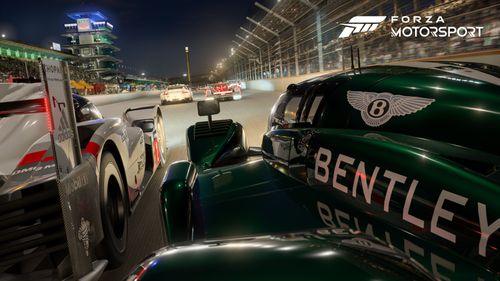 An onboard-style view from above the rear left wheel of the 2003 #7 Team Bentley Speed 8 as it races down the start finish straight at Indianapolis Motor Speedway with the 2017 #2 Porsche Team 919 Hybrid just to the left of the Bentley and slightly ahead.