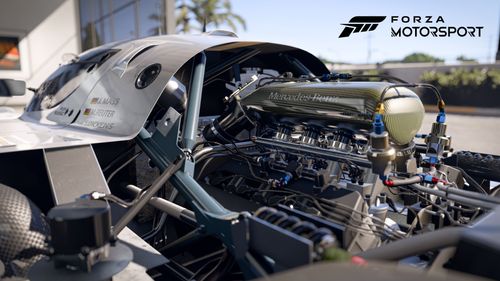 The engine of a Sauber C9 race car.  The grey car itself is sitting just inside a building that appears to have large glass garage-style doors. The view is zoomed in on the engine of the car so that intricate parts are clearly visible.