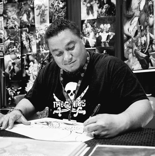 A black-and-white photo of a person wearing a black t-shirt with a pirate skull and swords on it. Behind where they are sitting, the wall is covered in comic-book-style illustrations. The person is intently focused on drawing on a paper in front of them.