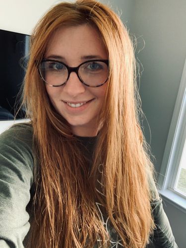 A white perosn with long orange/red hair smiles at the camera. They are wearing a gray sweater and glasses with large textured frames.